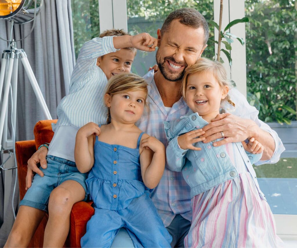 Sam's son, Brando sticking his finger in his dad's ear, while the twins sit on one knee each